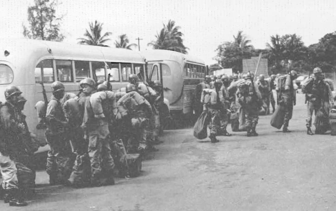 Soldiers with duffel bags boarding buses beneath palm trees